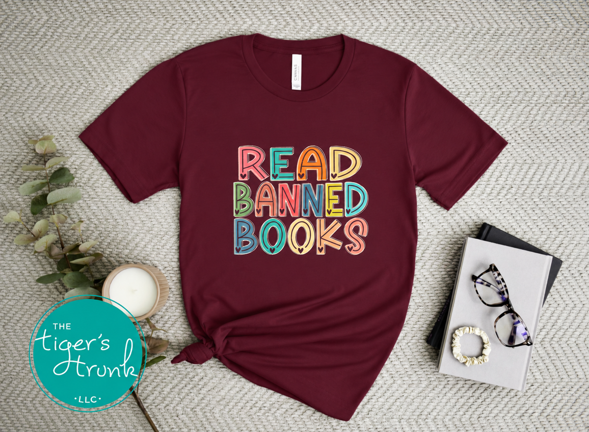 A maroon short-sleeve graphic T-shirt laid flat on a textured beige surface, with the phrase 'READ BANNED BOOKS' printed in colorful bubble letters. A small knot is tied at the bottom left hem.
