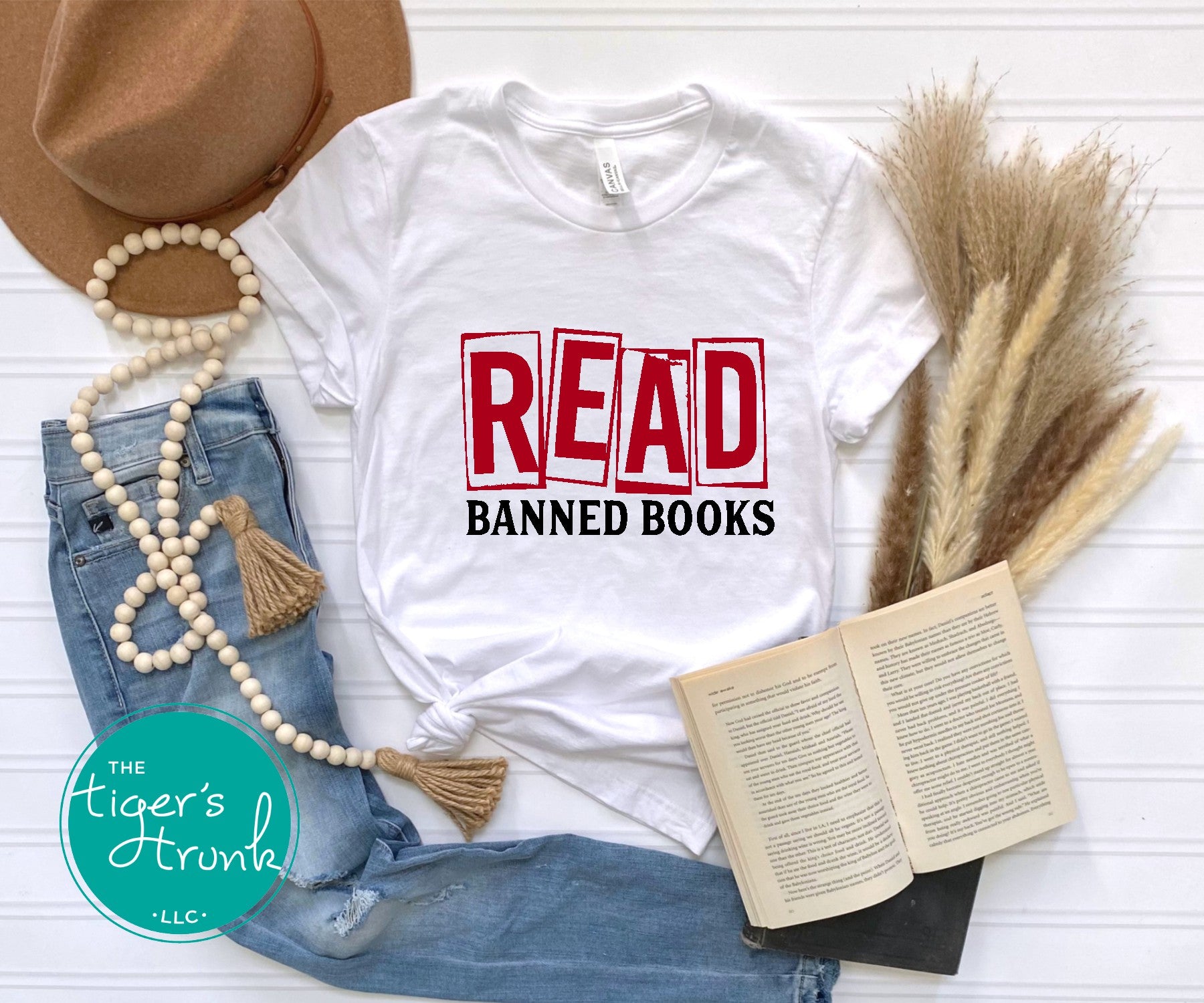 White t-shirt with 'READ BANNED BOOKS' text, surrounded by a hat, book, and decorative items on a white surface.