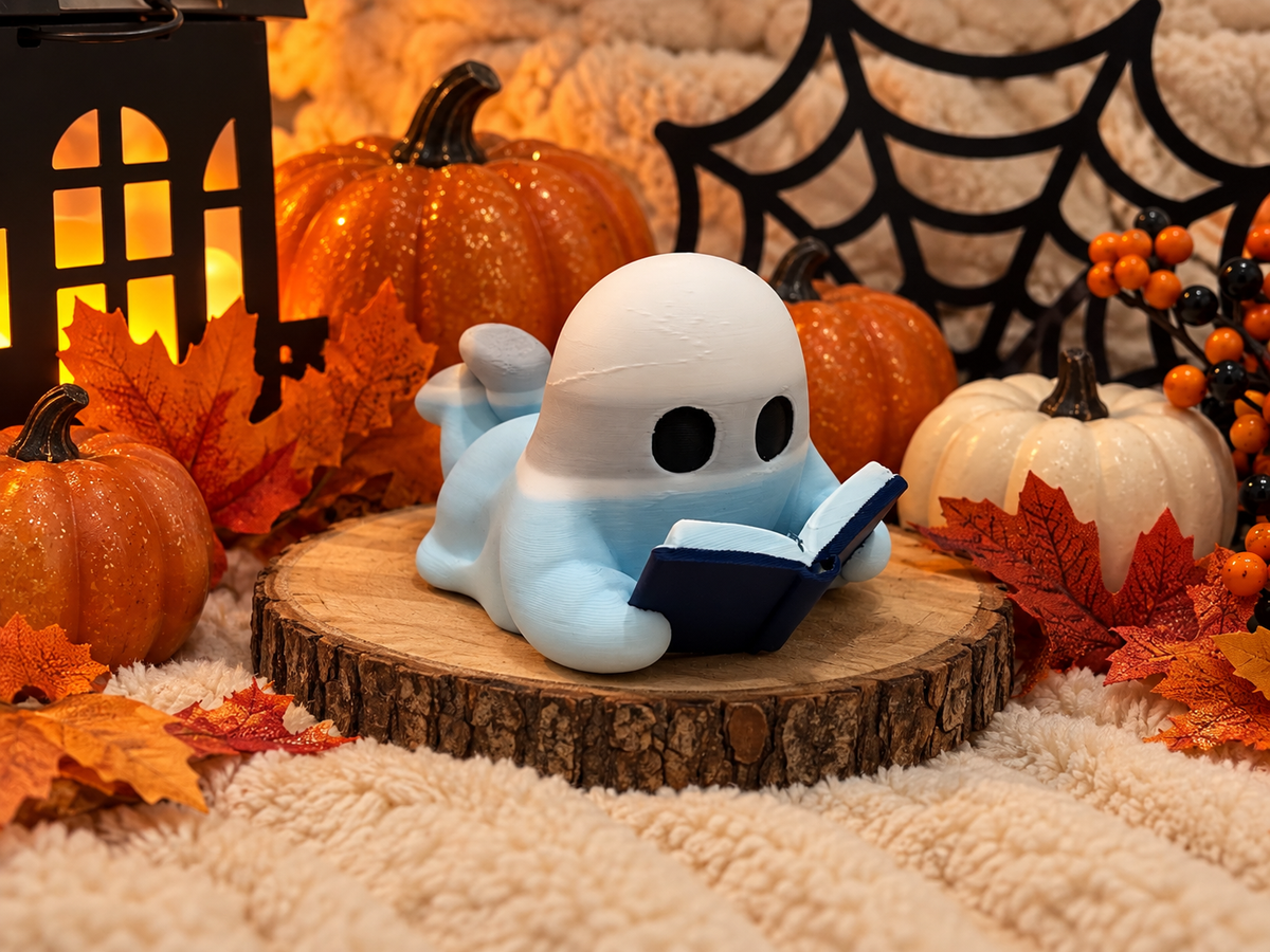 Cute white ghost figurine with black eyes reading a navy blue book, displayed among Halloween pumpkins and autumn decorations on a wooden riser