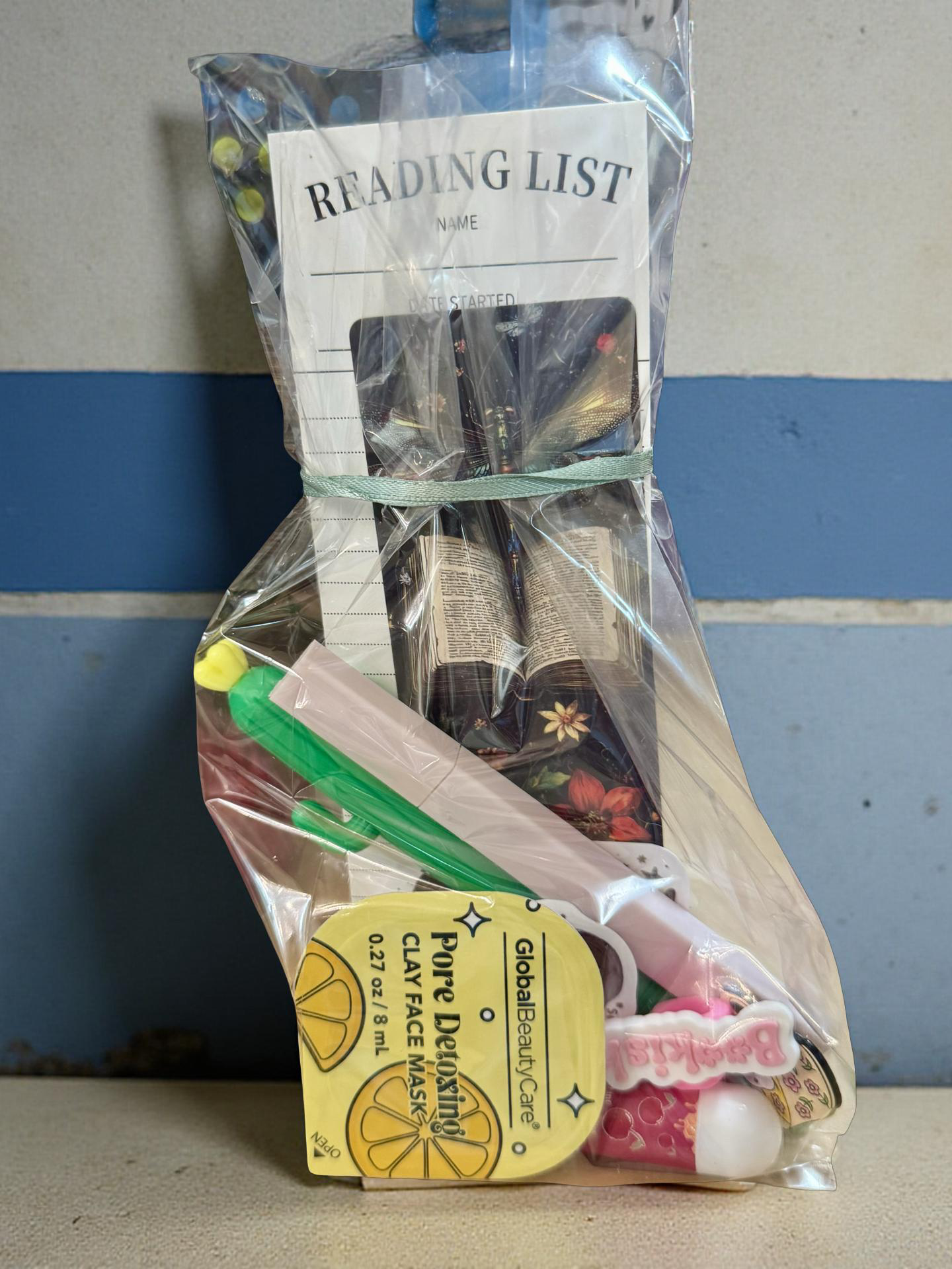 Goody bag filled with reading-themed items including a reading list planner, bookmark, highlighter, pen, stickers, enamel pin, chapstick, nail file, and face mask inside clear wrapping.