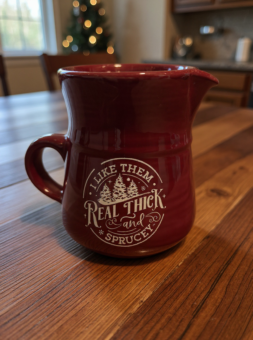 red ceramic pitcher-style mug with Real Thick and Sprucey design on wooden table