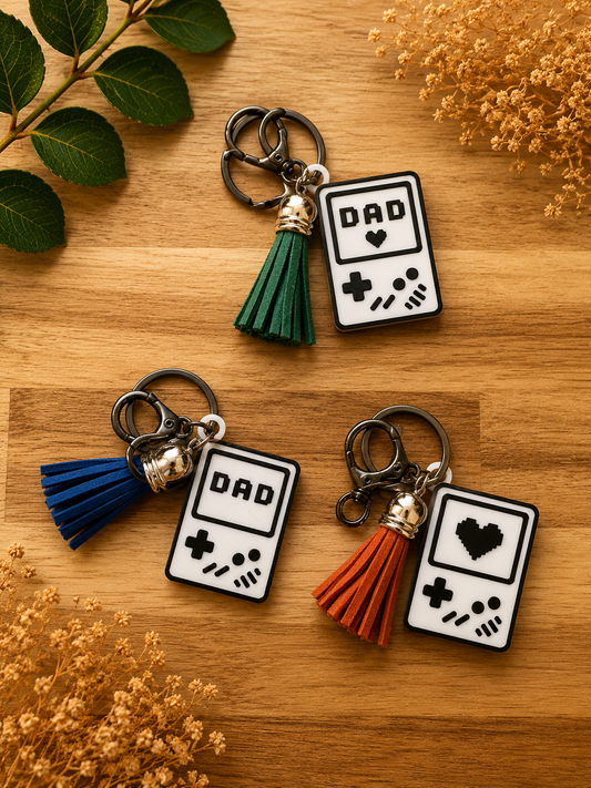 Set of retro gamer dad handheld console keychains with blue, green, and orange tassels arranged on wooden background