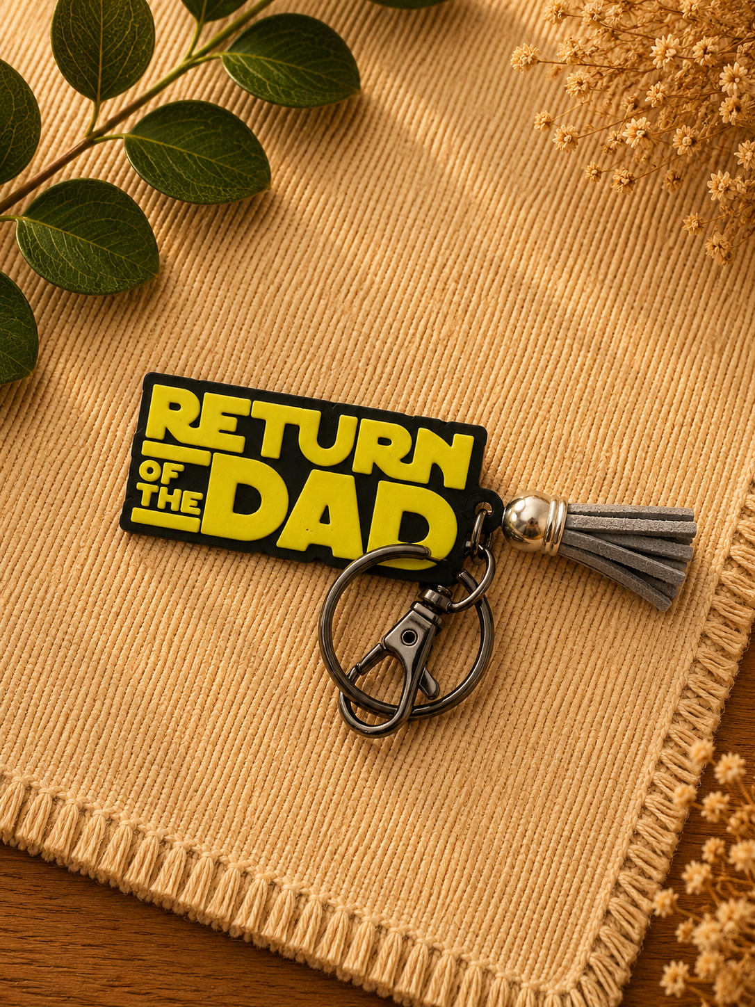 Black and yellow Return of the Dad 3D printed keychain with gray tassel displayed on woven neutral fabric