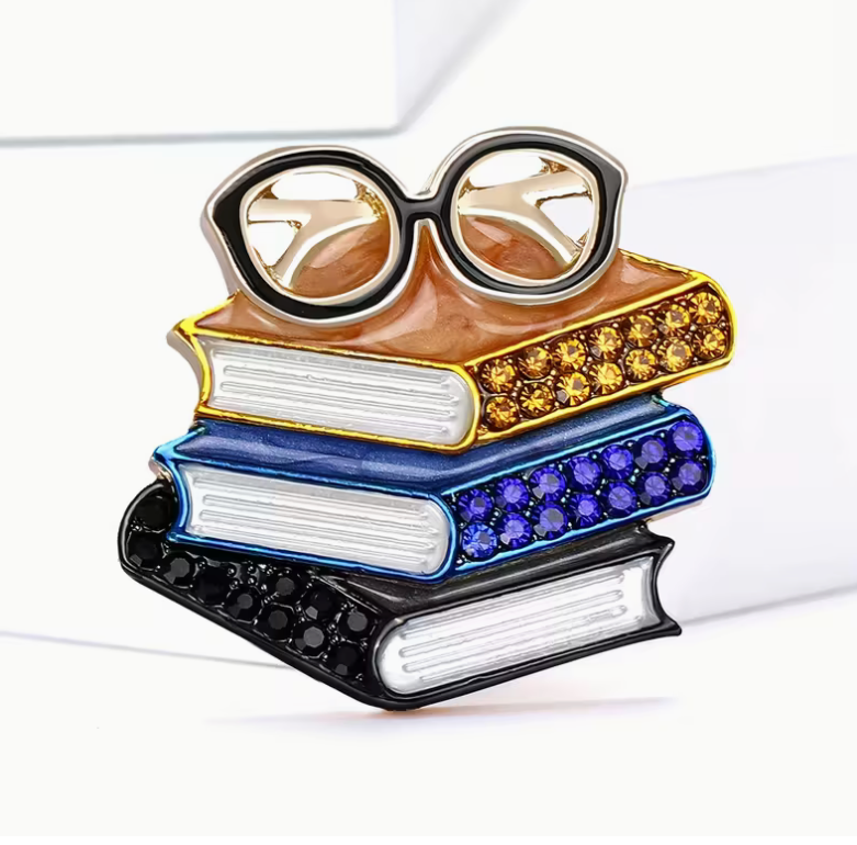 A decorative book-and-glasses accessory shaped like a stack of four books topped with eyeglasses. The books are colored black, blue, white, and gold, with rows of rhinestones on the top book and eyeglass frames.