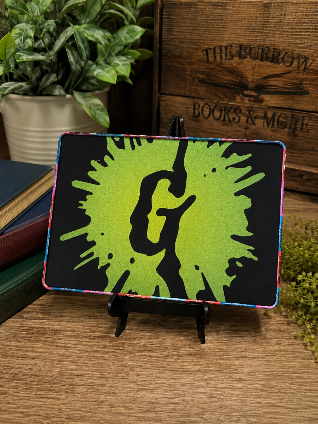 A rectangular black plaque or coaster with a bright lime green paint splatter design and a large black letter 'G' in the center, displayed on a small black stand on a wooden table with books and a potted plant in the background.