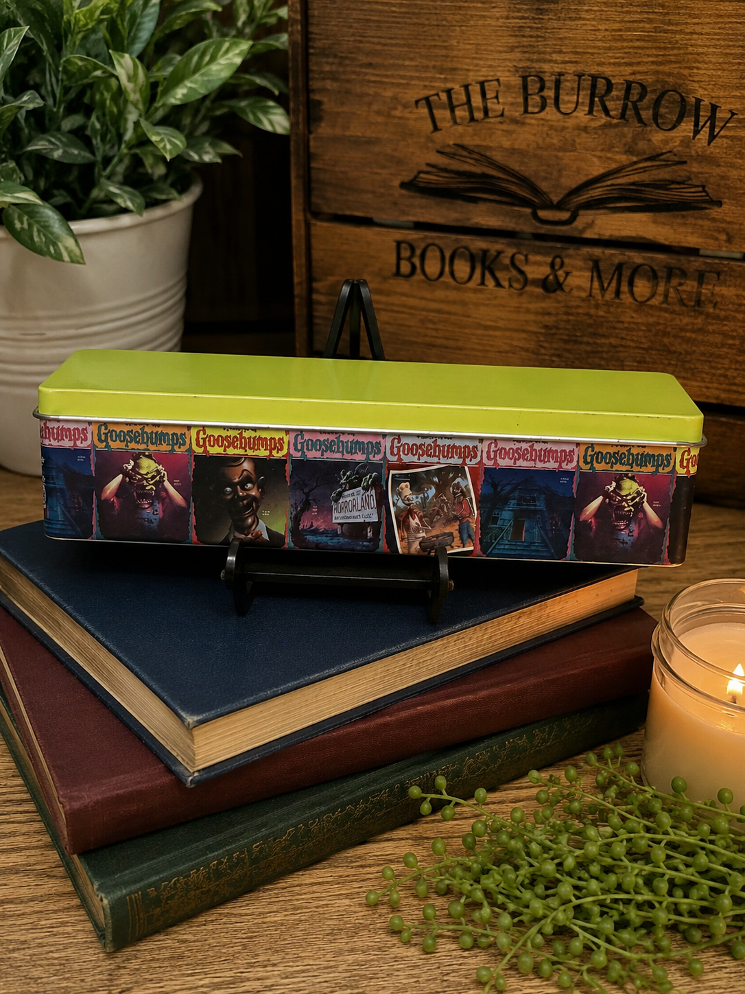 A hardcover book with a lime green top and a colorful illustrated spine, displayed on top of stacked books on a wooden table with a plant, candle, and wooden crate in the background.