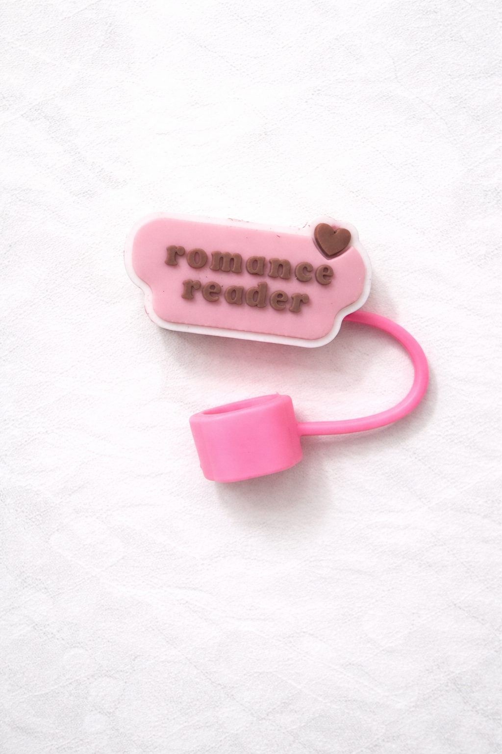 Pink silicone straw topper with raised lettering that says romance reader and a small heart detail, attached to a flexible loop for reusable tumbler straw.