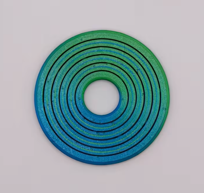 Flat round 3D printed fidget spinner in green and blue tones showing layered concentric ring design on a white background.