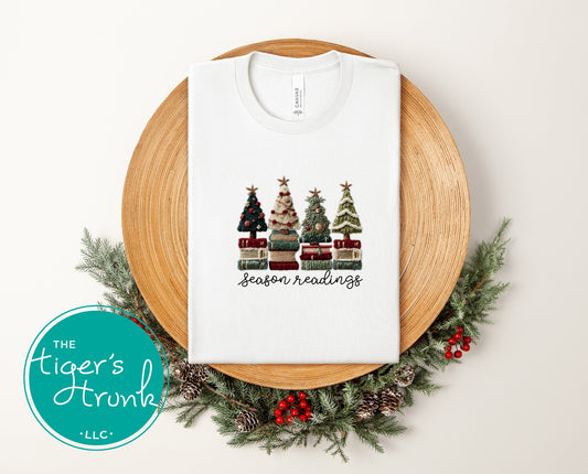 White t-shirt folded on a wooden tray with book-stack Christmas tree design and the phrase season readings, styled with holiday greenery.