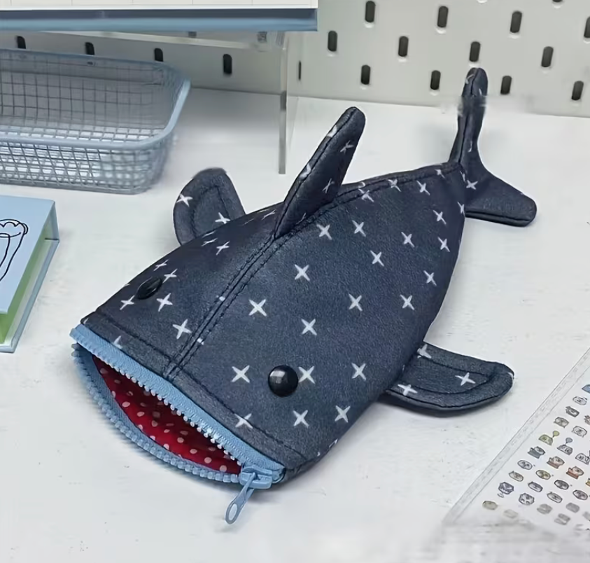 Gray shark-shaped canvas pencil case with zipper closure, white star pattern, fins, tail, and red polka-dot lining.