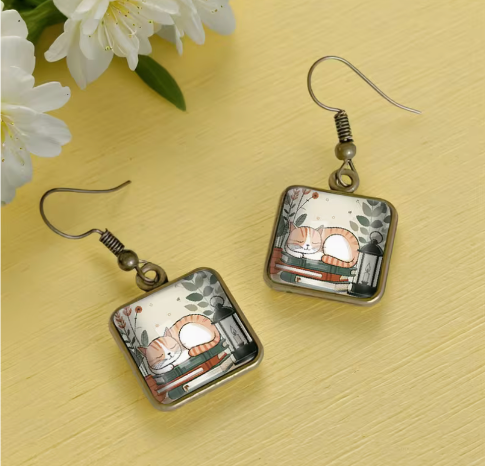 Square glass dangle earrings featuring an orange and white sleeping cat curled up on a stack of books