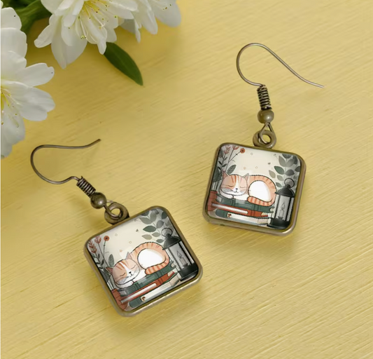 Square glass dangle earrings featuring an orange and white sleeping cat curled up on a stack of books