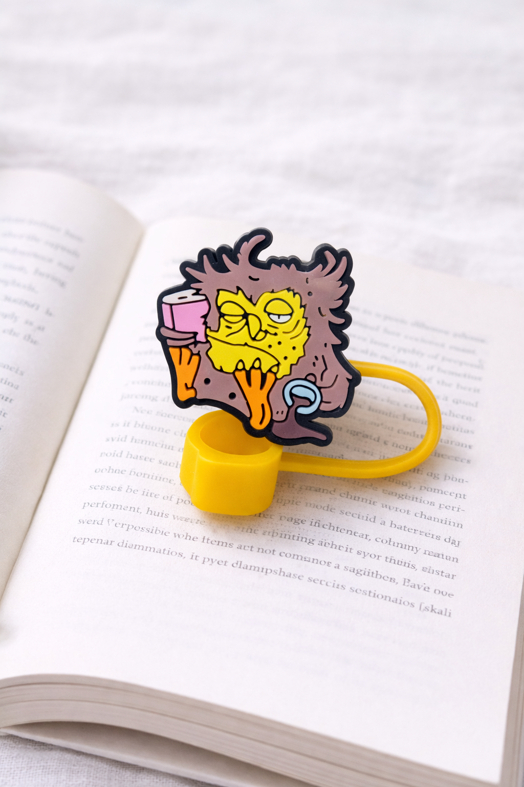 Cartoon sleepy book lover silicone straw topper with yellow loop resting on an open book page background.