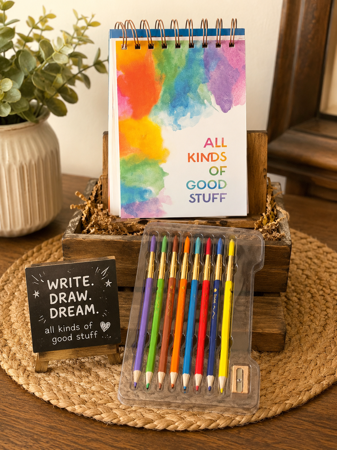 A stationery gift set displayed on a woven placemat in a home setting. The set includes a spiral-bound notepad with a colorful watercolor cover, a small black sign reading 'WRITE.