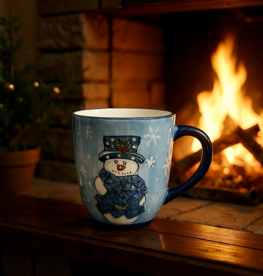 Blue ceramic mug with a painted snowman design sitting on a wooden ledge in front of a warm fireplace.