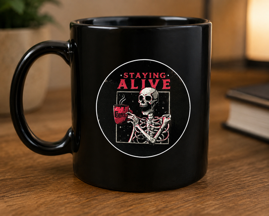 Black ceramic coffee mug featuring a Staying Alive skeleton sticker design with a skeleton holding a coffee mug, displayed on a wooden tabletop with books in the background.