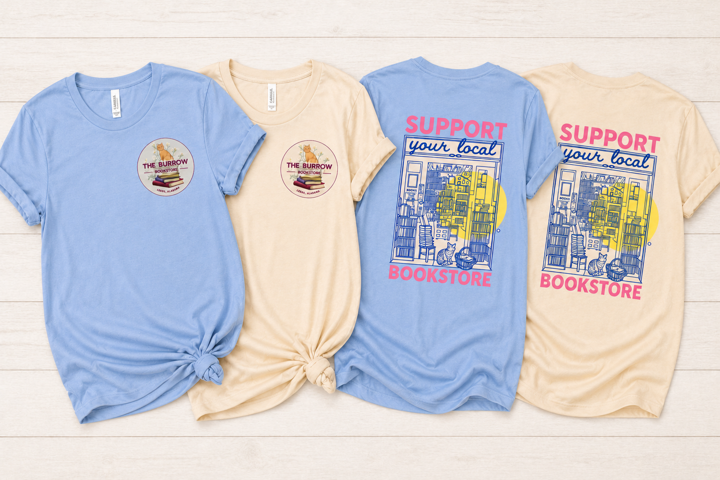 Four graphic t-shirts in light blue and cream are laid flat on a white wood background, showing both the front and back designs. The shirts feature a circular 'The Rainbow Bookstore' logo on the front chest and a larger 'SUPPORT your local BOOKSTORE' graphic with a colorful bookshelf illustration on the back.