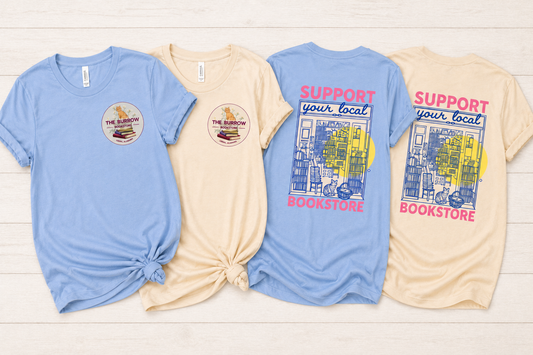 Four graphic t-shirts in light blue and cream are laid flat on a white wood background, showing both the front and back designs. The shirts feature a circular 'The Rainbow Bookstore' logo on the front chest and a larger 'SUPPORT your local BOOKSTORE' graphic with a colorful bookshelf illustration on the back.