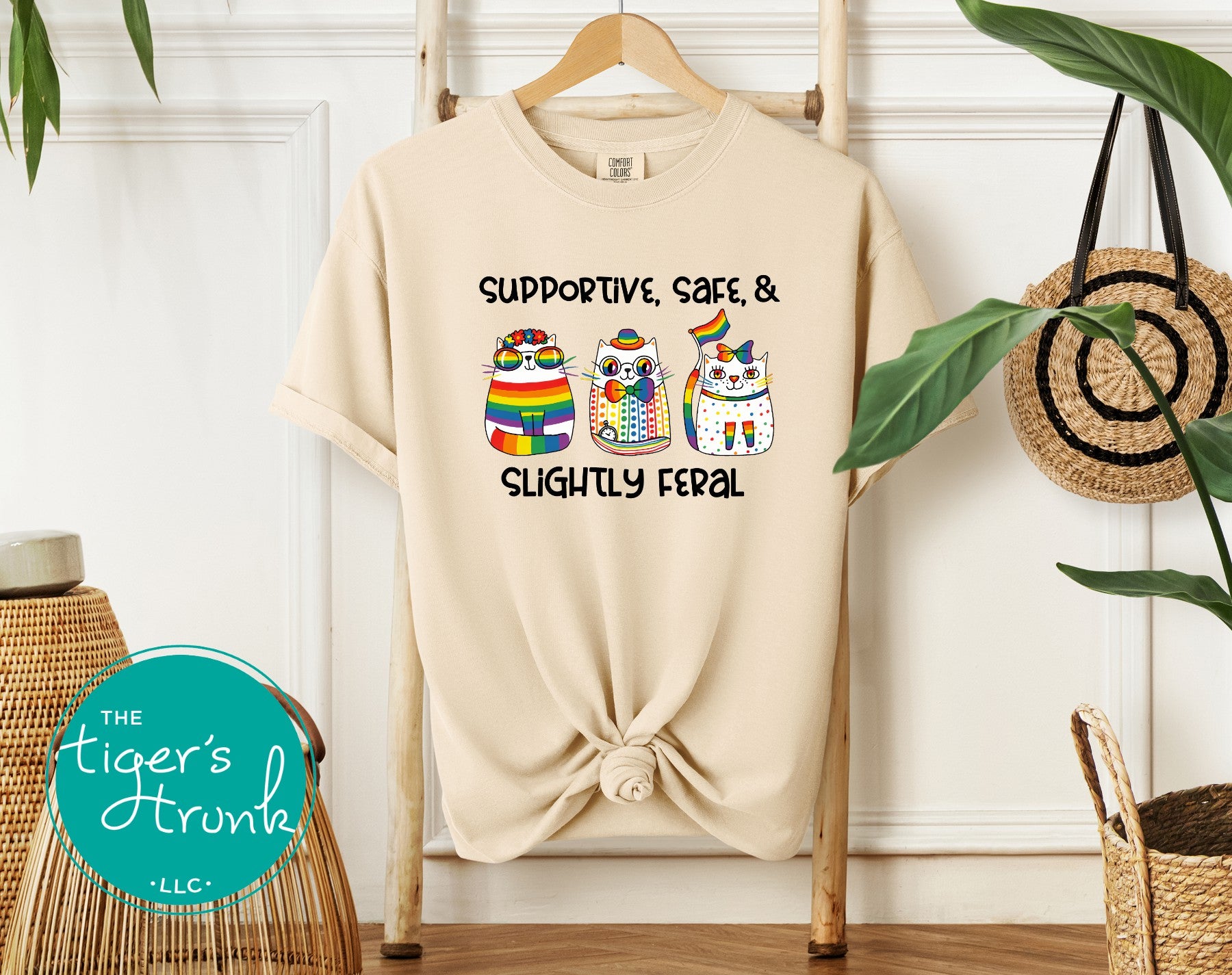 Beige t-shirt with rainbow cats and text on a hanger against a white wall.