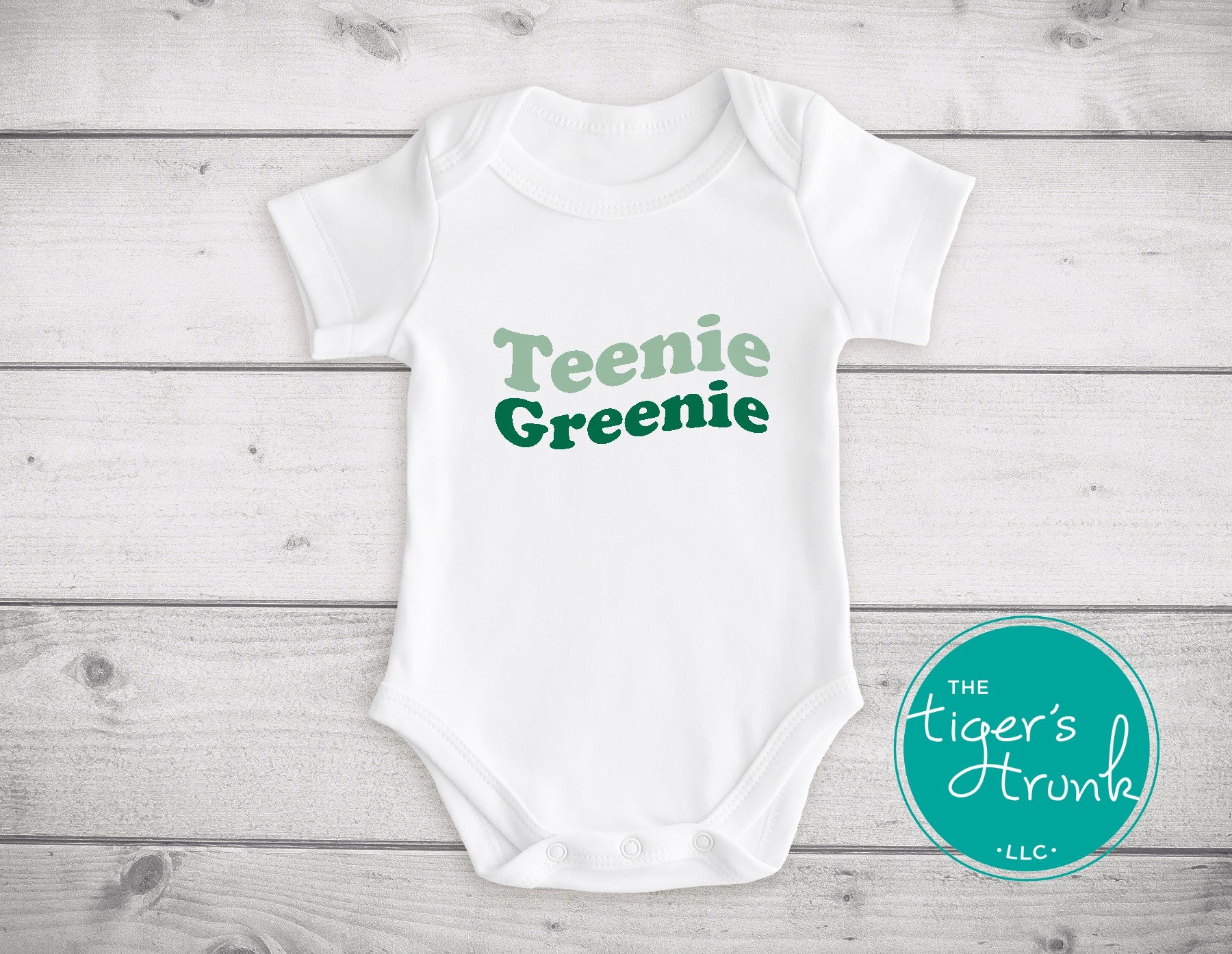 White baby onesie with 'Teenie Greenie' text on a wooden surface, featuring the brand 'The Tiger's Trunk'.