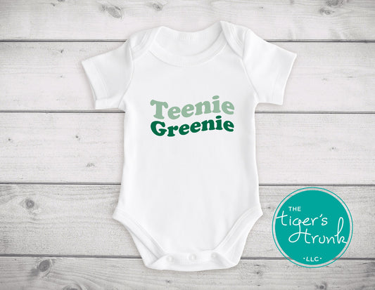 White baby onesie with 'Teenie Greenie' text on a wooden surface, featuring the brand 'The Tiger's Trunk'.