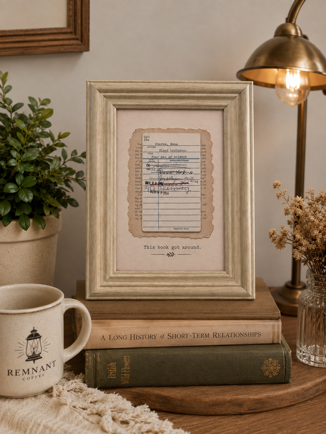 A framed vintage-style print featuring a lined script page with handwritten text and the caption 'THIS BOOK GOT AROUND.' displayed on a wooden table, surrounded by a mug, books, a plant, dried flowers, and a lamp.