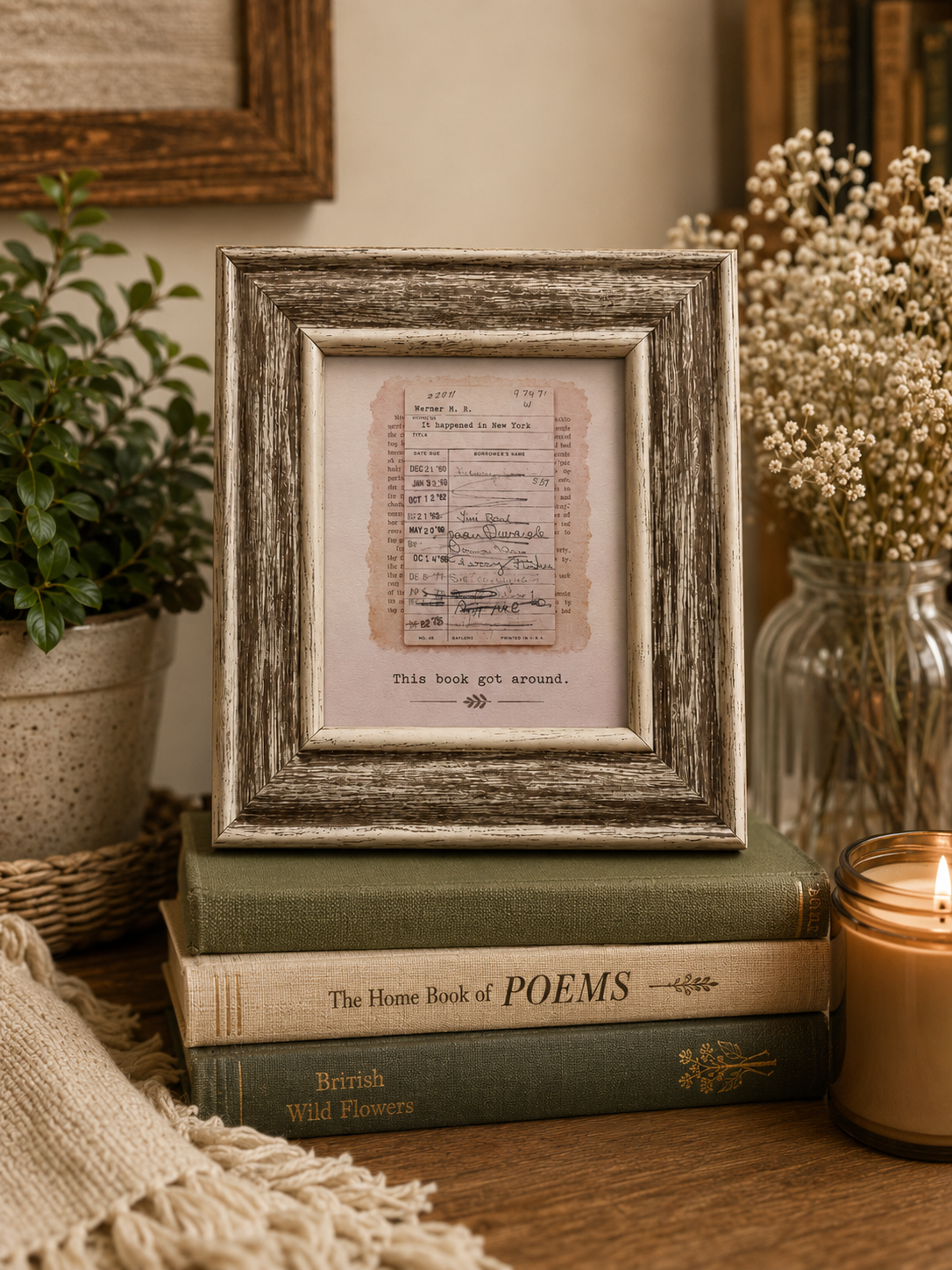 A rustic distressed wood picture frame displaying vintage handwritten musical notation on aged paper, styled on a stack of books on a wooden table with a lit candle, potted plant, and dried flowers in the background.