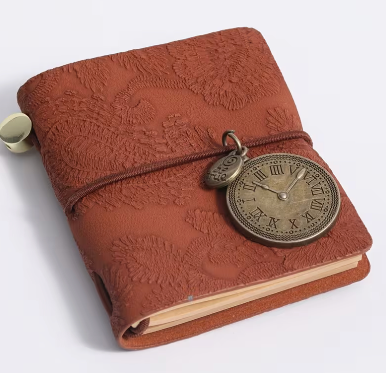 A brown embossed leather journal with a wrap-around elastic strap and a vintage-style metal pocket watch charm, shown on a clean white background.
