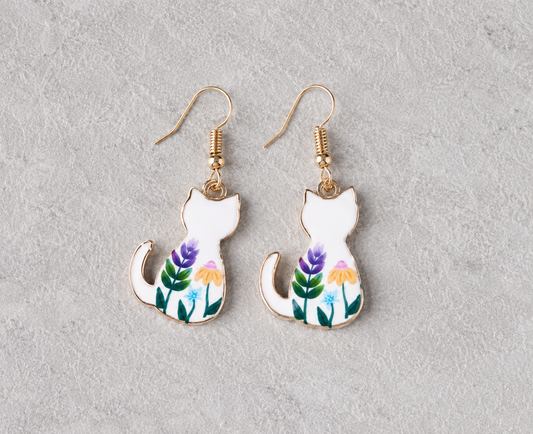 White cat pendant earrings with colorful floral enamel artwork and gold-tone hooks displayed on a neutral textured background