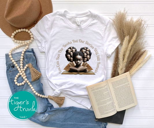 White t-shirt with graphic design featuring a child reading a book, surrounded by denim jeans, an open book, a hat, and decorative elements on a light wooden surface.
