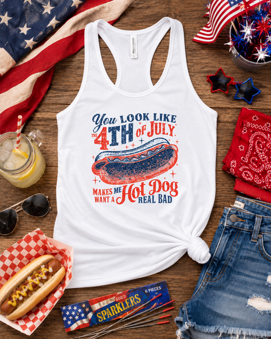 White racerback tank top with a patriotic 4th of July hot dog graphic, styled in a flat lay on a wooden surface with an American flag, denim shorts, sunglasses, a hot dog, a drink, and red, white, and blue party accessories.